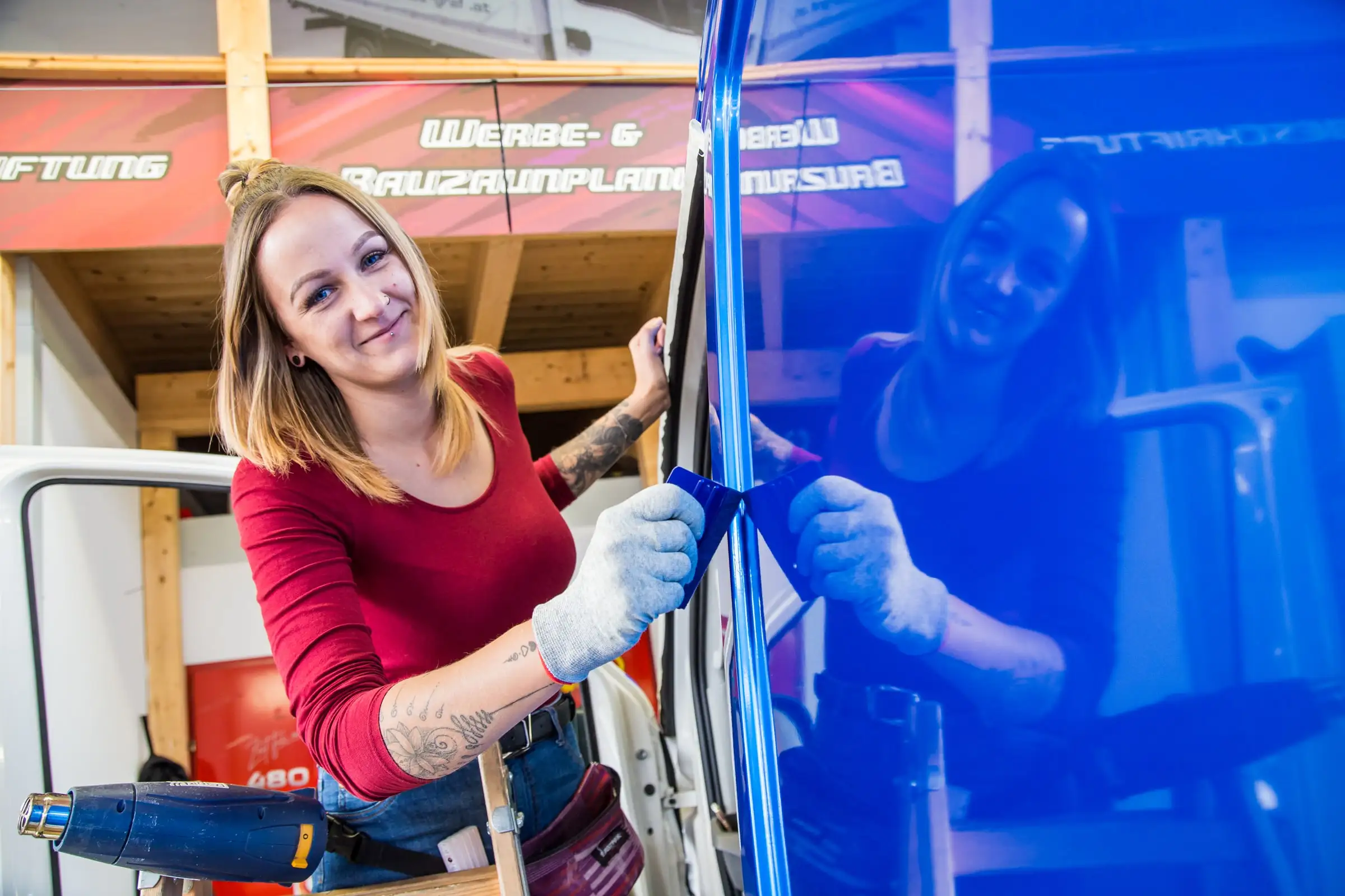 Mitarbeiterin von Schriften Tarmann zieht mit Rakel und Heißluftpistole eine blaue Folie an einem Transporter an, im Hintergrund die Werkstatt in Villach