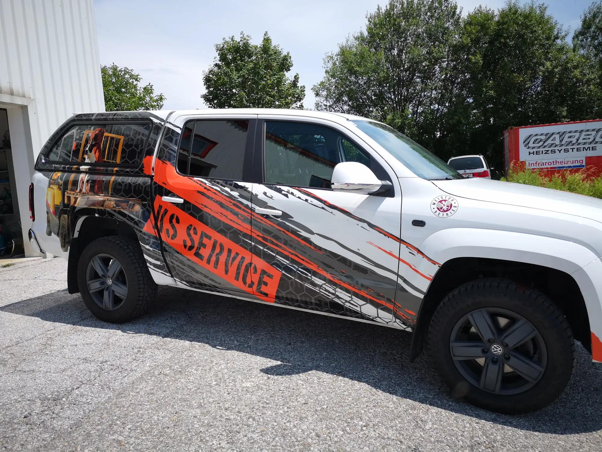 Weißer Pick-up mit Doppelkabine und vollflächiger Folierung in Orange und Grau vor der Halle in Villach