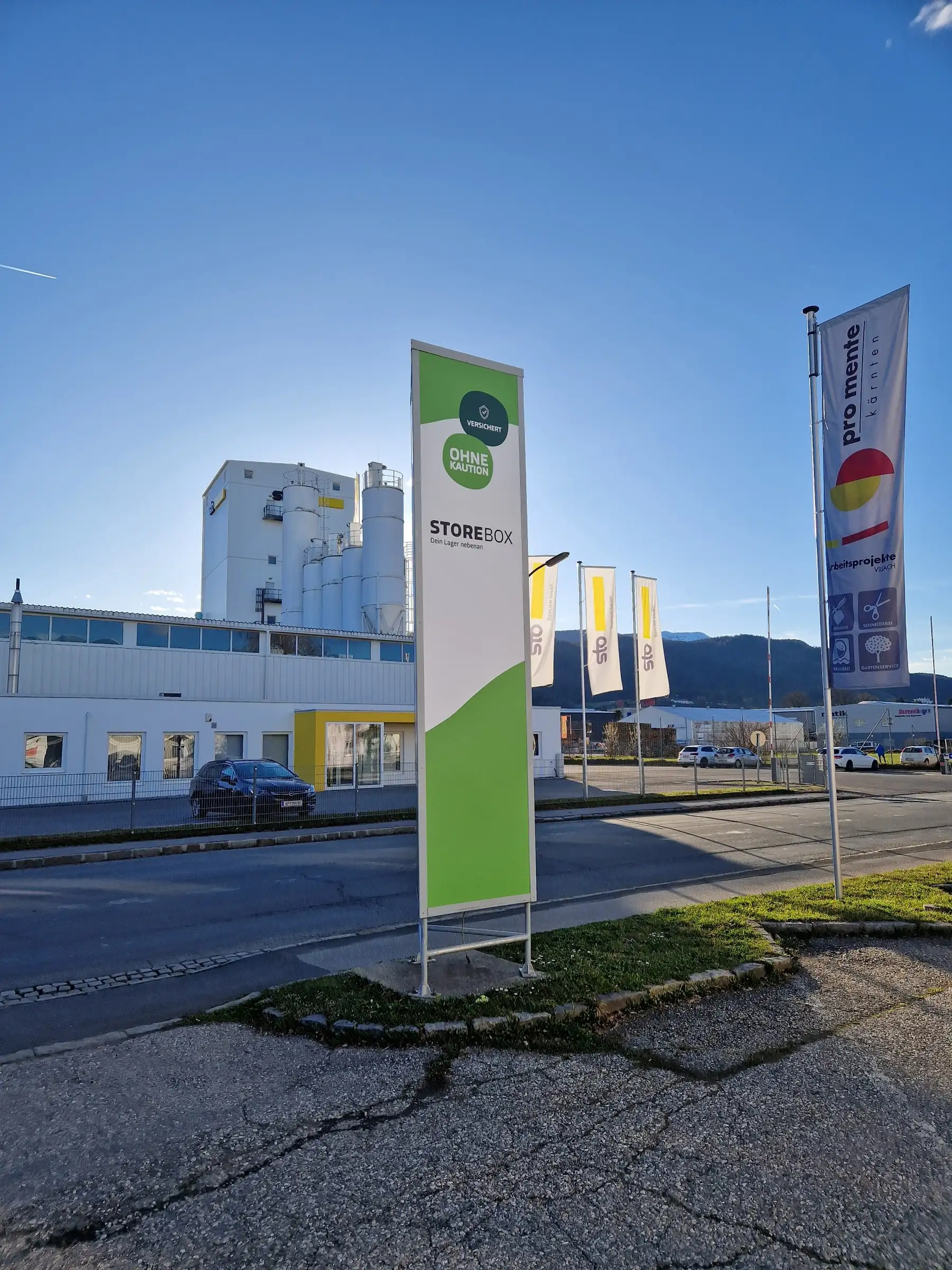 Weiß-grüner Werbepylon an der Straße, daneben mehrere bedruckte Werbefahnen vor einem Industriegebäude