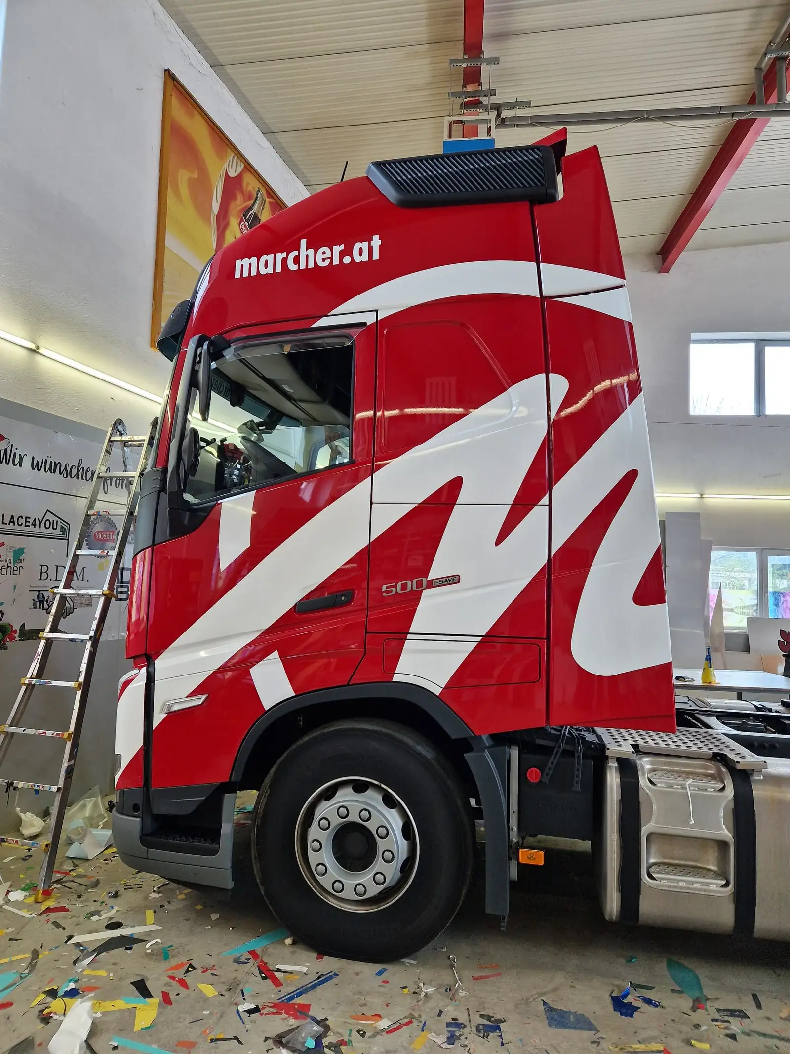 Rote Sattelzugmaschine mit weißem Schwungdekor, daneben Leiter und Folienreste während der Verklebung